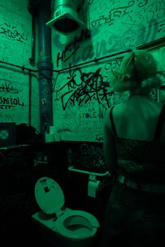 a woman standing in front of a toilet with graffiti on the wall behind her and green light