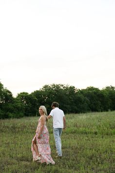 a man and woman are walking through the grass together in an open field with trees behind them