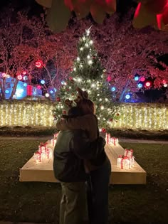 two people standing in front of a lit christmas tree with presents on the table under it