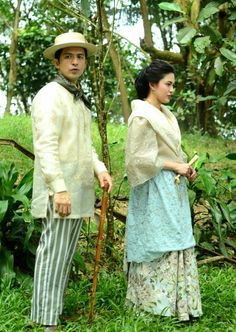 a man and woman standing next to each other in the grass with trees behind them