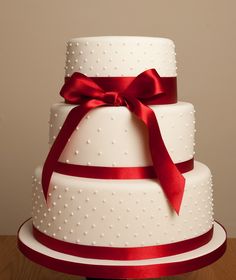 a three tiered wedding cake with red ribbon and bow on top, sitting on a wooden table