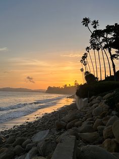 the sun is setting over the ocean with palm trees in the foreground and rocks on the shore