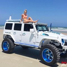 a woman sitting on top of a white jeep at the beach with blue rims