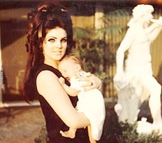 a woman holding a baby in her arms near a fountain with a statue behind her