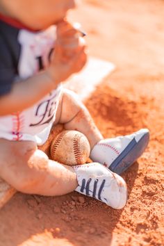 a little boy sitting on top of a baseball field holding a bat and ball in his hand