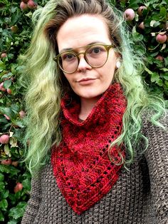 a woman with green hair wearing glasses and a red knitted scarf in front of bushes