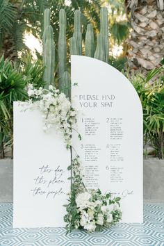 a wedding seating chart with white flowers and greenery on the side, next to a cactus