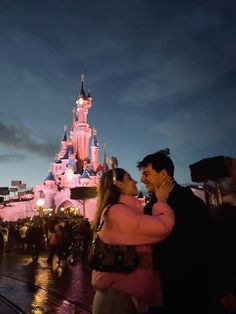 a man and woman standing in front of a castle at night with their arms around each other