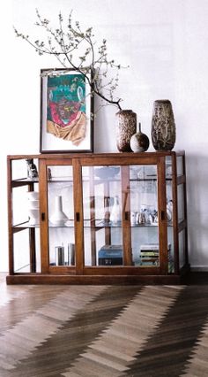 a wooden cabinet with glass doors and vases on top, in front of a white wall