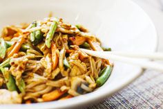 a white bowl filled with noodles and veggies next to chopsticks on a table