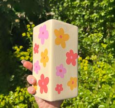 a hand holding up a wooden block with flowers on it in front of some bushes