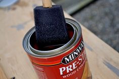 a paint can with a brush in it sitting on top of a wooden table next to a piece of wood