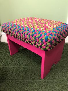 a pink bench with multicolored knitted seat cushion on green carpet in front of white wall