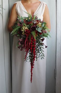 a woman in a white dress holding a red and green wedding bouquet with greenery