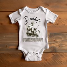 Rapunzie's charming baby onesie that celebrates the special bond between fathers and their little ones. Made from 100% cotton, this onesie is designed for ultimate softness and comfort, ensuring a delightful experience for your baby. The highlight of this onesie is its heartwarming graphic illustration featuring a father fishing with his young son. The beautiful scene captures the joy and connection shared between a dad and his little fishing buddy. Accompanying the illustration is the adorable Father's Day Gift Cotton Onesie, Father's Day Family Matching Onesie, Cotton Onesie For Father's Day, Short Sleeve Onesie For Father's Day Gift, Father's Day Casual Short Sleeve Onesie, Baby Fish, A Father, Baby Bodysuit, Graphic Illustration