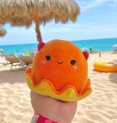 a hand holding an orange stuffed animal on the beach with umbrellas in the background