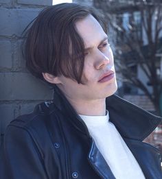 a man leaning against a brick wall wearing a black jacket and white t - shirt