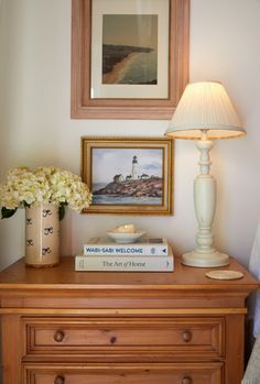 a nightstand with books and flowers on it next to a painting, lamp and pictures