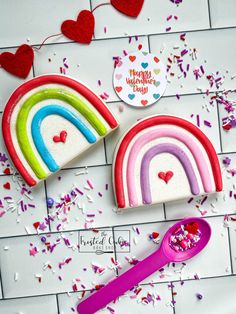 two rainbow shaped cookies sitting on top of a white tile floor next to a pink spoon