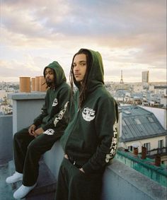 two young men sitting on top of a roof wearing green sweatshirts and sweatpants