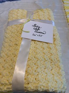 a yellow crocheted blanket with a white ribbon tied around it and a baby gift tag on top