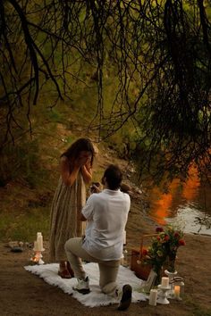 a man kneeling down next to a woman on top of a blanket near water and trees