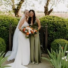 two women standing next to each other in front of bushes and trees with flowers on them