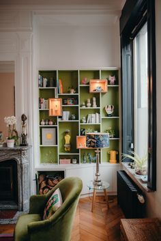 a green chair sitting in front of a window next to a fire place and bookshelf