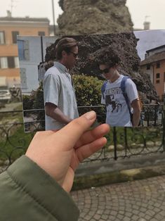 a hand holding up a photo of two men in front of a stone building and fence