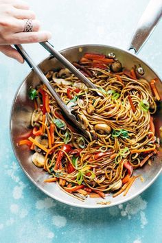 two hands holding tongs over a bowl of stir fry noodles with carrots and mushrooms