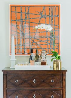 a dresser with bottles and glasses on it in front of an orange wall art piece