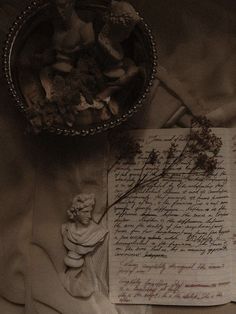 an old letter and statue next to a bowl of dried flowers on a cloth covered table