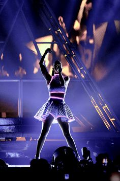 a woman is performing on stage with her arms in the air and lights behind her