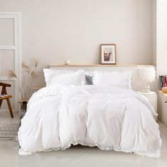 a bed with white comforter and pillows in a room next to a chair, table and pictures on the wall