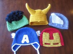 four crocheted hats are arranged on a wooden table, one with an iron man hat and the other with a spiderman hat