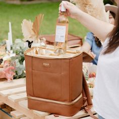 a woman is holding a bottle of wine in front of a bag on a table