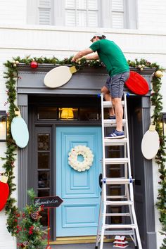 a man on a ladder painting the front door of a house with wreaths and christmas decorations