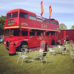 a red double decker bus parked on top of a grass covered field next to tables and chairs