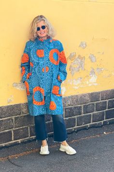 a woman standing in front of a yellow wall wearing sunglasses and a blue dress with orange circles on it