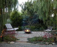 a fire pit in the middle of a garden surrounded by trees and bushes with chairs around it