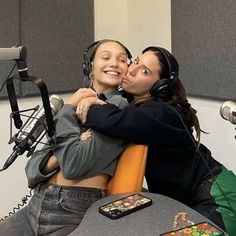 two women sitting in front of microphones with headphones on and one is hugging the other