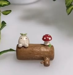 two small figurines sitting on top of a piece of wood next to plants