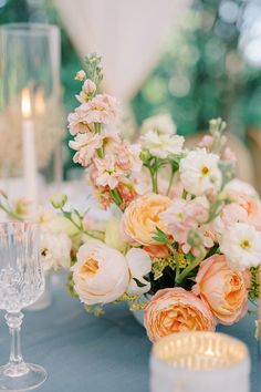 an arrangement of flowers and candles on a blue table cloth with glassware in the foreground