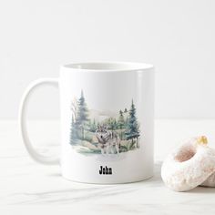 a white coffee mug with a wolf and trees on it next to a donut