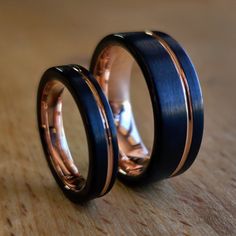 two wedding bands with black and rose gold inlays are sitting on a wooden surface