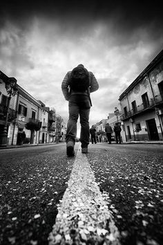 a person walking down the middle of an empty street with buildings in the background and clouds overhead