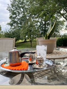 a glass table topped with a tea pot next to a metal tray filled with cups and saucers