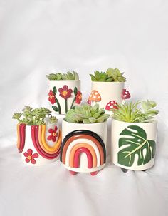 four ceramic pots with plants in them on a white cloth covered surface, one has a rainbow and the other has flowers