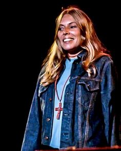 a woman standing in front of a black background wearing a denim jacket and cross necklace