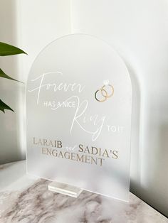 a clear acrylic sign with two wedding rings on it sitting next to a potted plant